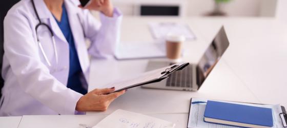 una donna in camice e con stetoscopio al collo è seduta alla scrviania e consulta il pc mentre parla al cellualre e tiene in mano una cartella di dcoumenti