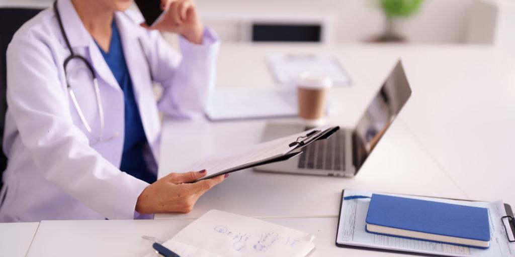 una donna in camice e con stetoscopio al collo è seduta alla scrviania e consulta il pc mentre parla al cellualre e tiene in mano una cartella di dcoumenti